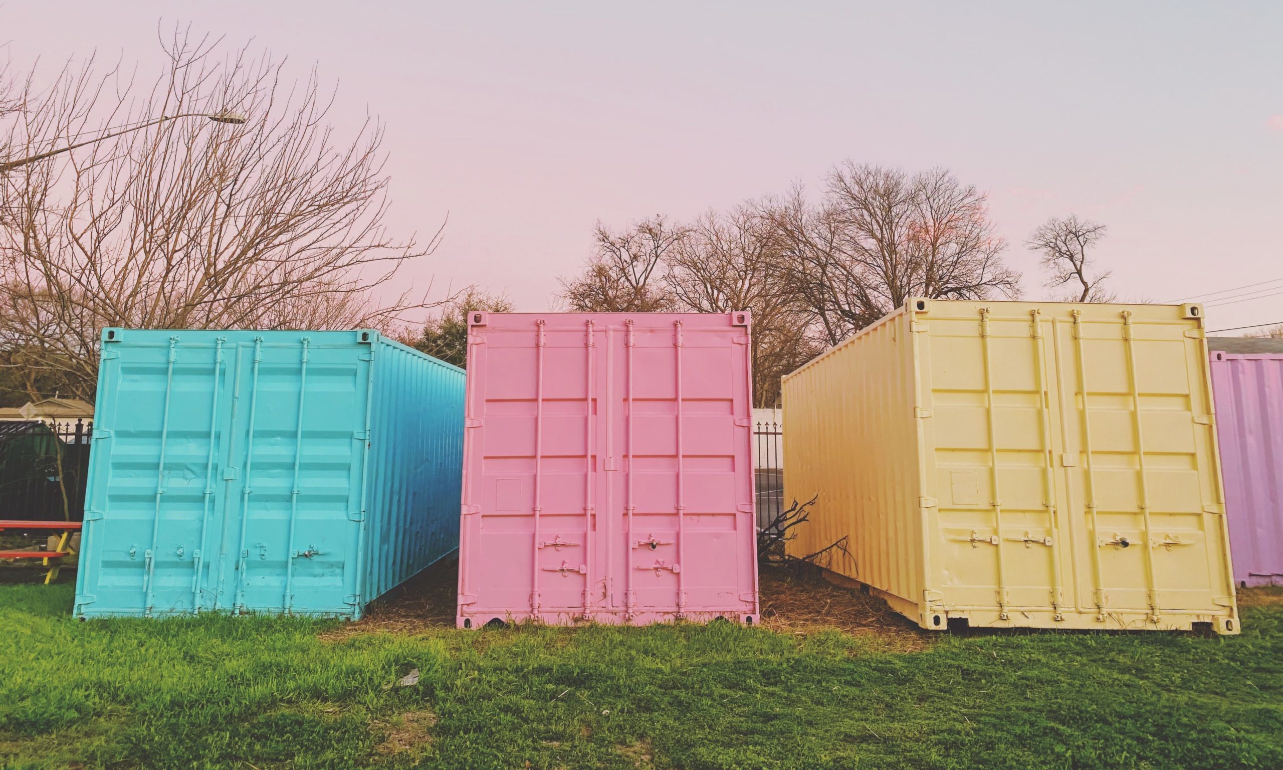 Trois conteneurs maritimes colorés (turquoise, rose, jaune) alignés sur une pelouse.